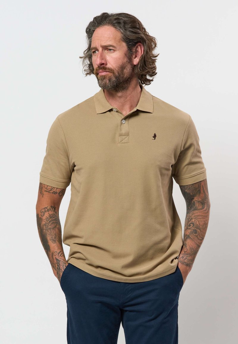 Mann mit schulterlangem Haar und Bart, der ein beiges Polohemd und marineblaue Hose trägt, Hände in den Taschen, schaut zur Seite, mit tätowierten Armen.