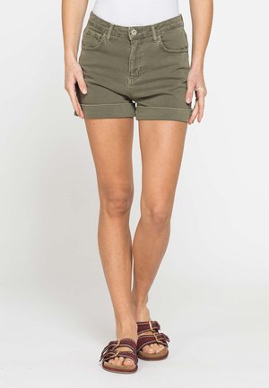 Persona che indossa pantaloncini verde oliva con orlo arrotolato, top bianco e sandali color granata con doppia fibbia, in piedi davanti a uno sfondo chiaro e uniforme.