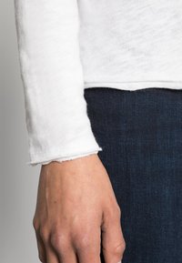 Camiseta blanca de manga larga con puños deshilachados y textura suave, combinada con tela de mezclilla oscura, que muestra un ajuste casual y un diseño simple.