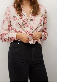 Blouse à manches longues, rose à motif floral avec boutons sur le devant ; tissu semblant léger avec une texture douce ; portée avec un jean noir taille haute.