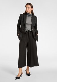 Schwarz gestreifter Blazer mit doppelreihigem Design, grauer Rollkragenpullover, schwarze weitgeschnittene Hose und schwarze Slingback-Absätze mit Schleifen.