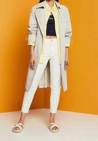 Žena oblečená v béžovém trenčkotě přes žluté tričko a námořnické crop top, bílé kalhoty a béžové sandály na oranžovém pozadí.