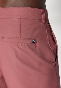 Rosa treningsshorts med teksturert stoff, med baklomme, beltestropper og en liten merketag på siden.