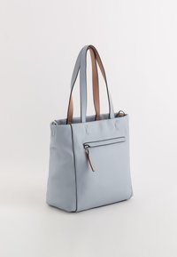 Lysblå læder tote bag med brune håndtag og en frontlomme med lynlås, der har en tan lædertrækklap. Glat tekstur med struktureret form.
