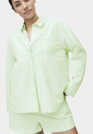 Kvinde iført lysegrøn og hvidstribet skjorte med knapper og matchende shorts, poserer med den ene hånd på hoften.