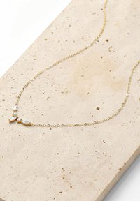 Goldene Kettenhalskette mit drei dekorativen Akzenten aus klaren Steinen, vor einer strukturierten, hellen Steinoberfläche.