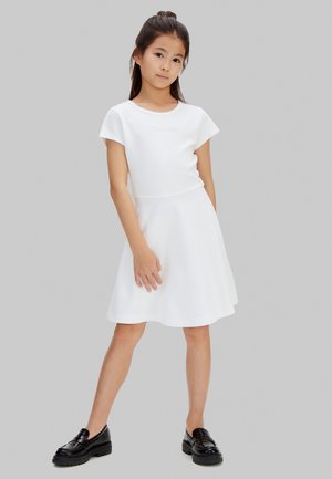 Ung pige stående iført en hvid kjole med korte ærmer og sorte loafers mod en ensfarvet grå baggrund.