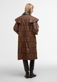 Langer brauner Brokatmantel mit einem kunstvollen orangefarbenen Blumenmuster, breitem Kragen und gepufften Ärmeln. Kombiniert mit dunklen kniehohen Lederstiefeln.