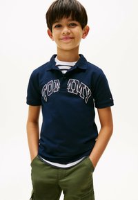 Marineblå polo t-shirt med "TOMMY" logo, korte ærmer og krave; båret over en hvid og stribet skjorte, parret med grønne cargo shorts.