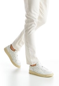 Sneaker bianche in pelle con dettagli perforati e una suola di gomma beige. Le sneaker presentano lacci e un design a bassa altezza. Indossate con pantaloni chiari.