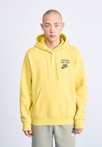 Sárga Nike kapucnis felső, elől zsebből, húzózsinórral a kapucnin, és "CSAK HIVATALOS HASZNÁLATRA" felirattal. Rendelkezik alap стандартos fazonnal és puha pamut anyaggal.