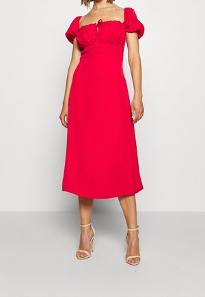 Kobieta ubrana w jasnoczerwoną, midi sukienkę odkrywającą ramiona z bufkami na rękawach i beżowe sandały na obcasie, stojąca na gładkim tle.