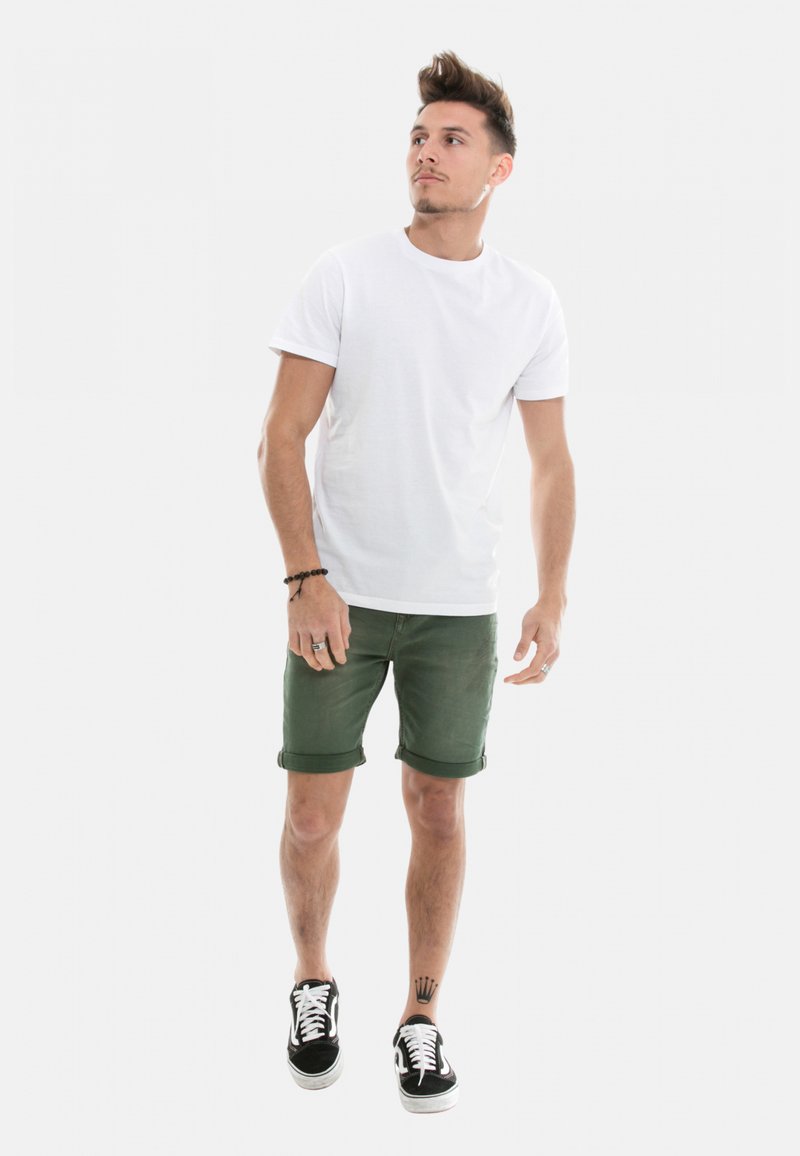 T-shirt en coton blanc, shorts en denim vert olive et baskets noir et blanc. L'ensemble présente des ourlets retroussés et un design décontracté.