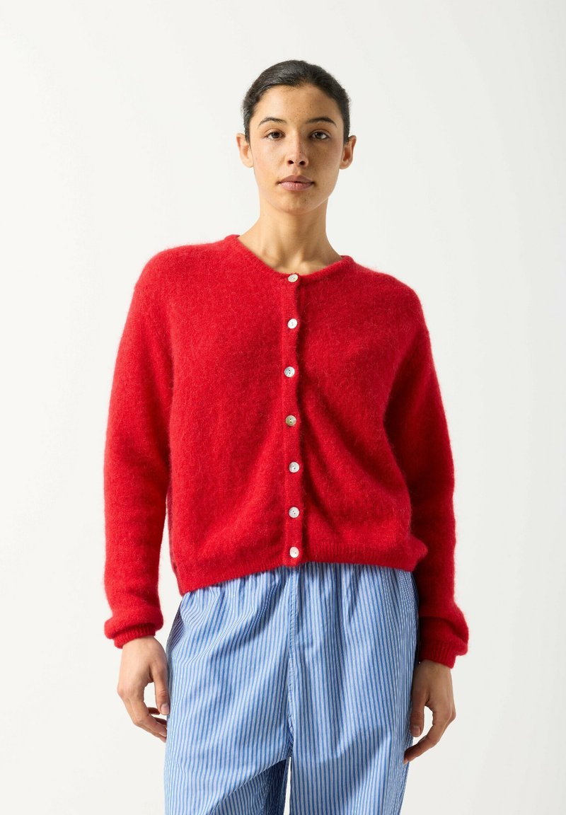 Žena v jasne červenom plyšovom svetri na zapínanie a vo voľných modro-bielych pruhovaných nohaviciach na jednoduchom svetlom pozadí.