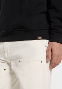 Felpa nera in cotone con orlo a coste, abbinata a pantaloni bianchi in tela con bottoni metallici argento e tasche minimaliste.