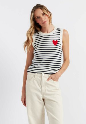 Top sans manches rayé en blanc et bleu marine, arborant une appliqué en forme de cœur rouge sur la poitrine. Associé à un pantalon beige clair.