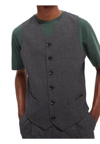 Uomo che indossa una camicia verde a maniche corte sotto un gilet grigio a scacchi con bottoni, pantaloni coordinati, mani in tasca.