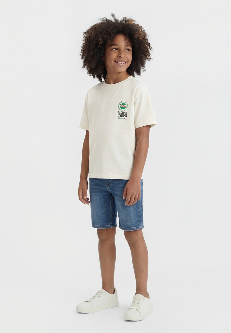 Enfant souriant aux cheveux bouclés portant un t-shirt blanc Castrol Racing, un short en jean et des baskets blanches, debout sur un fond blanc.