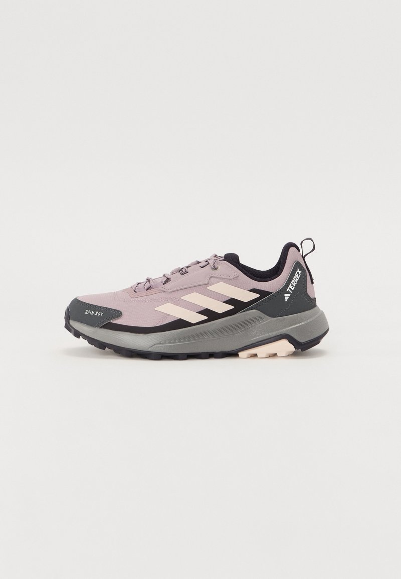 Chaussure de trail Adidas Terrex mauve clair et noire avec des bandes beiges, une semelle épaisse et une languette de tirage sur fond uni.