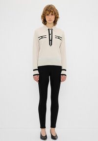 Maglia a maniche lunghe color crema in maglia con dettagli neri, colletto e bottoni. Indossata con leggings neri e scarpe basse. Design minimalista.