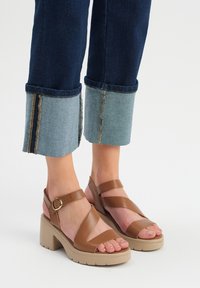 Brązowe skórzane sandały z szerokimi krzyżującymi się paskami, z klamrą na kostce i masywną beżową platformą. W zestawie z podwiniętymi ciemnymi dżinsami.