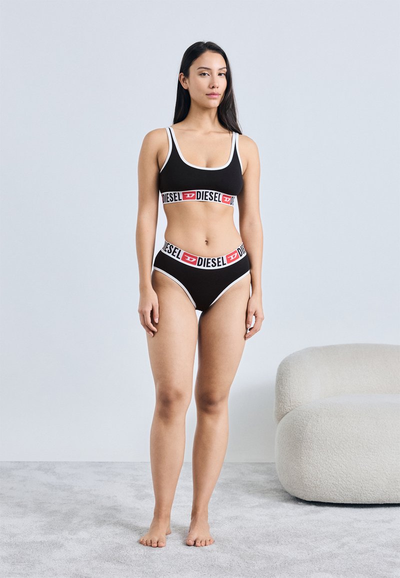 Schwarzes Crop-Top und Bikini-Set mit weißem Rand, mit einem auffälligen rot-weißen Logoband über der Taille. Weiche Stoffstruktur, minimalistisches Design.