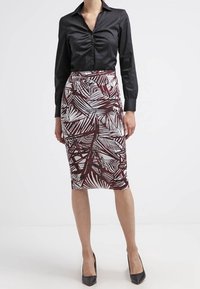 Femme portant un blouse noire cintrée avec un devant froncé et une jupe midi avec des motifs abstraits bruns et blancs, associée à des talons noirs.