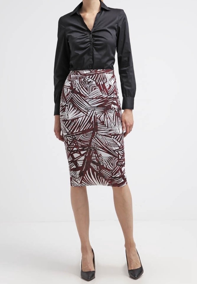 Femme portant un blouse noire cintrée avec un devant froncé et une jupe midi avec des motifs abstraits bruns et blancs, associée à des talons noirs.