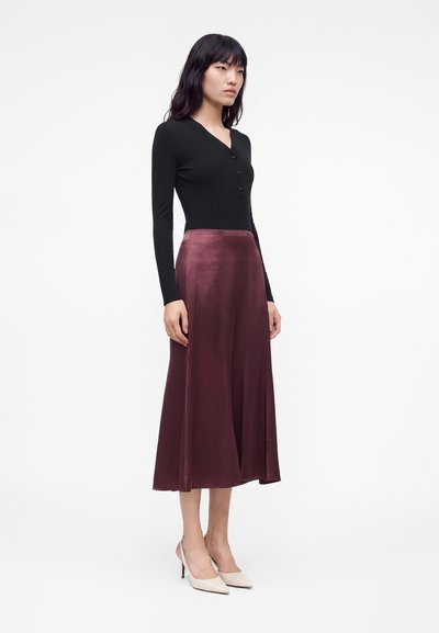 Femme aux longs cheveux foncés portant un haut noir à manches longues, une jupe midi en satin bordeaux et des talons hauts pointus de couleur claire.