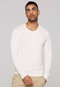 Langärmliges V-Neck-Hemd aus hellbeigem Stoff mit feiner Struktur. Die Ärmel sind eng anliegend und das Hemd ist leicht locker geschnitten.