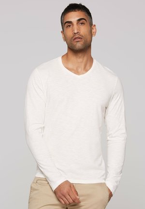 Langärmliges V-Neck-Hemd aus hellbeigem Stoff mit feiner Struktur. Die Ärmel sind eng anliegend und das Hemd ist leicht locker geschnitten.