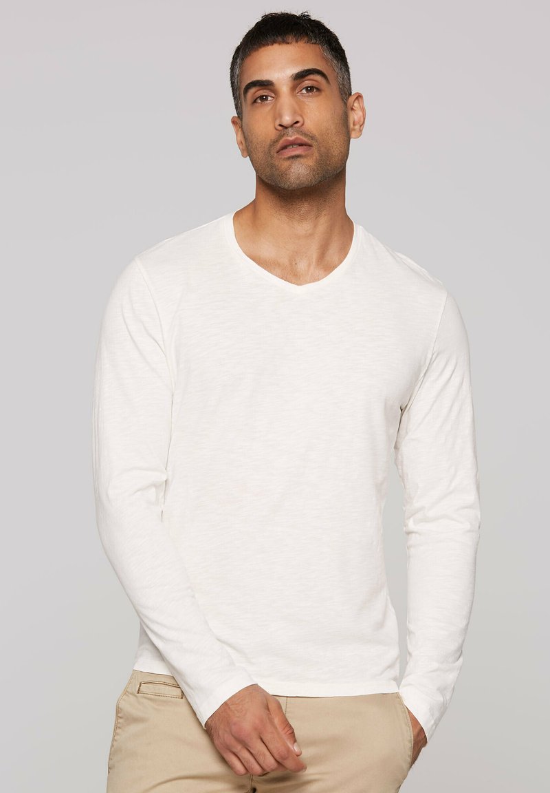 Langärmliges V-Neck-Hemd aus hellbeigem Stoff mit feiner Struktur. Die Ärmel sind eng anliegend und das Hemd ist leicht locker geschnitten.