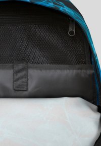 Rygsäck med ett blått mönster, svart mesh-inredning och en slät vit bottenfack. Dragkedjestängning och tygöglor för grepp.