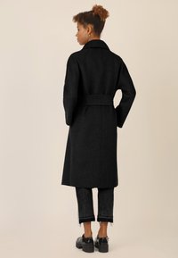 Zwarte wollen jas met een ceintuur in de taille, een brede kraag en lange mouwen, gecombineerd met donkere cropped jeans en chunky zwarte schoenen.