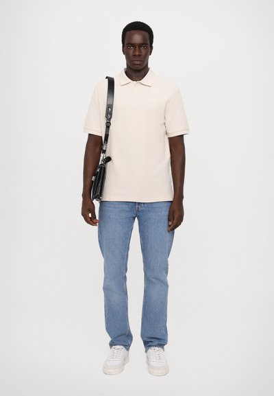 Lys beige poloskjorte, blå jeans, hvite sneakers. Modellen står med en svart crossbody-veske, som har en skulderstropp og sølvfarget detaljer.