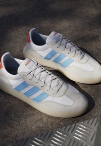 Par hvide Adidas sneakers med lyseblå striber, beige ruskindsdetaljer og gennemsigtige såler placeret på en tekstureret overflade.