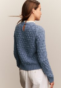 Mujer vistiendo un suéter de punto azul texturizado con un cierre de botón en forma de llave en la parte de atrás y pantalones blancos, de perfil contra un fondo simple.