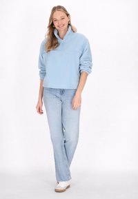 Pullover in pile azzurro chiaro con colletto zip, maniche lunghe e vestibilità rilassata, abbinato a jeans di denim chiaro e sneaker bianche.