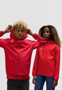 Red lightweight jackets with hoods, featuring zippered front, elastic cuffs, and logo detail on the chest. Smooth, water-resistant material.