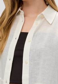 En blond person som bär en genomskinlig vit skjorta med knappar över en svart topp, vilket visar kragen och övre bröstområdet.