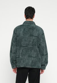 Σκούρο πράσινο μπουφάν από φλις με υφή σχεδίου, μανσέτες με κουμπιά και γιακά. Μαλακό, ζεστό υλικό με άνετη εφαρμογή.