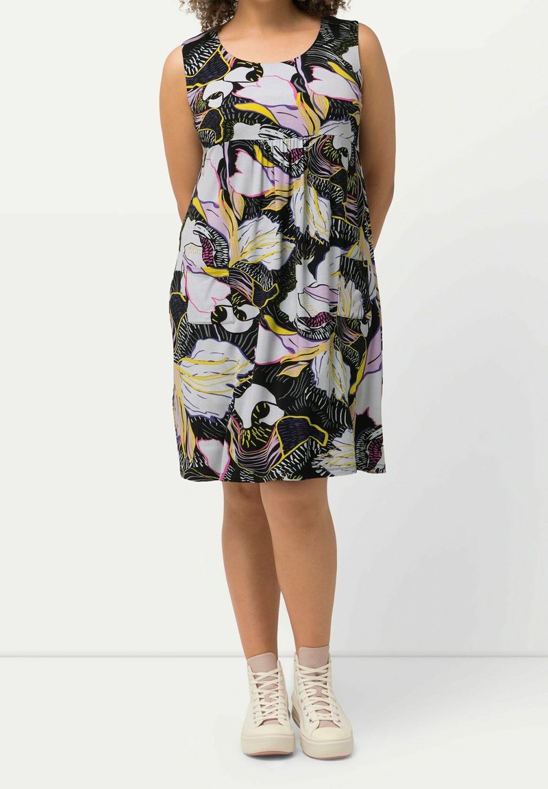 Femme portant une robe sans manches longueur genou avec un imprimé floral abstrait en noir, blanc, jaune et violet, associée à des baskets montantes blanches.