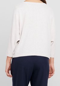 Haut ample beige clair à manches trois-quarts, tissu texturé, ourlet arrière arrondi, assorti à un pantalon bleu marine.