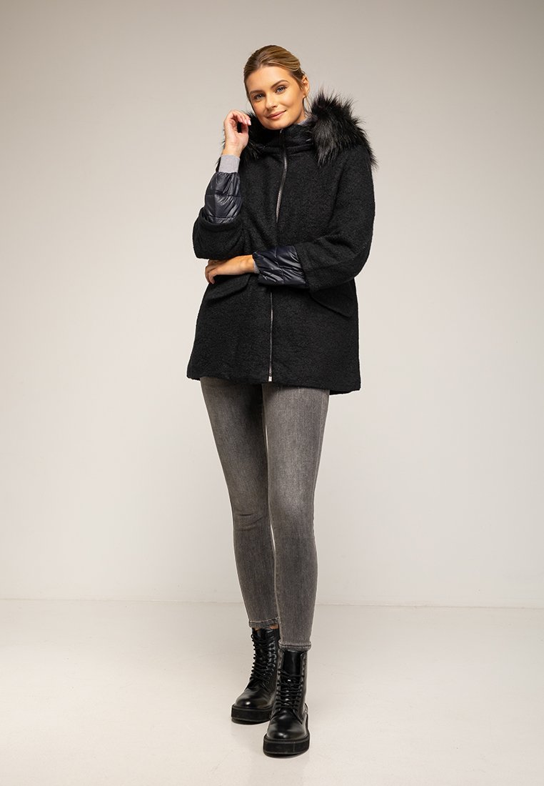 Czarny płaszcz z kapturem z sztucznego futra, teksturowany materiał, zamek błyskawiczny i pikowane rękawy. W zestawie z szarymi legginsami i czarnymi butami na sznurówki.