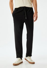 Sorte joggebukser med justerbar talje, tætsiddende ben og manchetter ved anklerne. Fremstillet af glat, blødt stof. Båret med hvide sneakers.