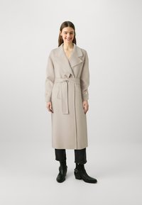 Cappotto lungo beige in lana con ampio colletto, cintura in vita annodata, tasche laterali e polsini con bottoni. Indossato sopra jeans neri a vita alta e stivaletti neri.