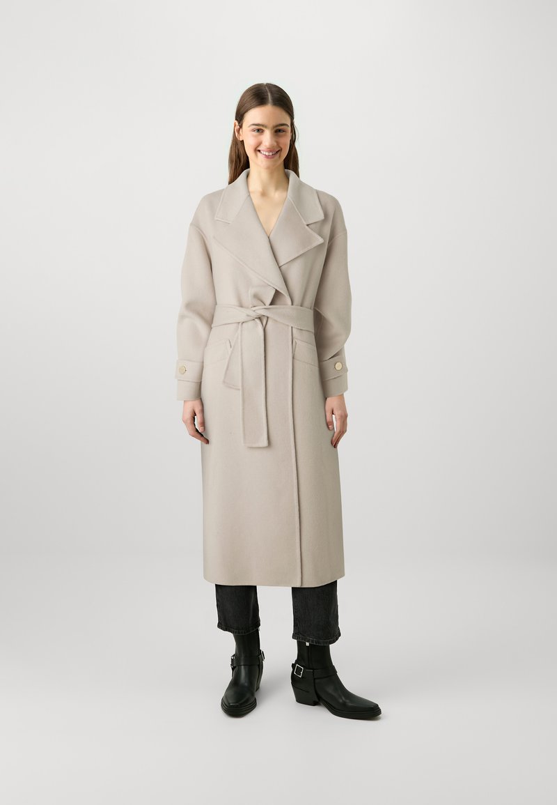 Cappotto lungo beige in lana con ampio colletto, cintura in vita annodata, tasche laterali e polsini con bottoni. Indossato sopra jeans neri a vita alta e stivaletti neri.