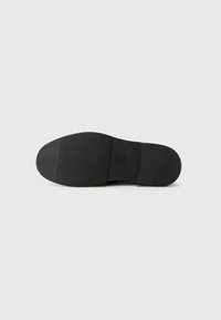 Semelle de chaussure noire avec une finition texturée, présentant une forme arrondie et des bords lisses, marquée d'un logo sur le côté.