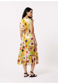 Blumenkleid in Midi-Länge mit Rüschensäumen, rosa Grundfarbe, kräftigen mehrfarbigen Blumenmustern, gestuftem Rock und Taillendetail. Getragen mit schwarzen Sandalen.