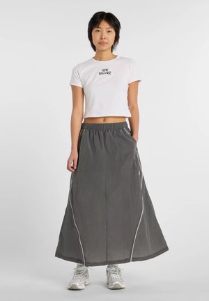 Mujer con camiseta blanca New Balance recortada, falda larga gris con ribete blanco y zapatillas blancas, de pie sobre un fondo liso.